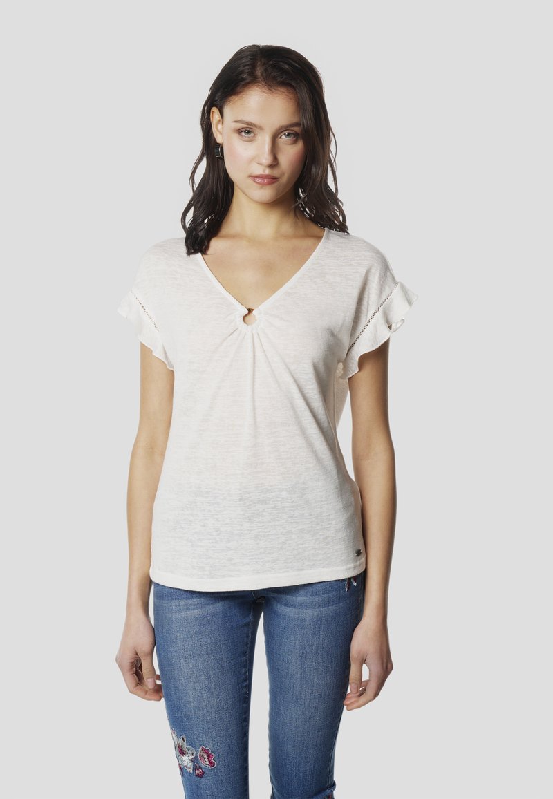 Haut blanc à col en V avec manches courtes à volants, doté d'un détail œillet au niveau du col. Porté avec un jean bleu, mettant en valeur une broderie florale.