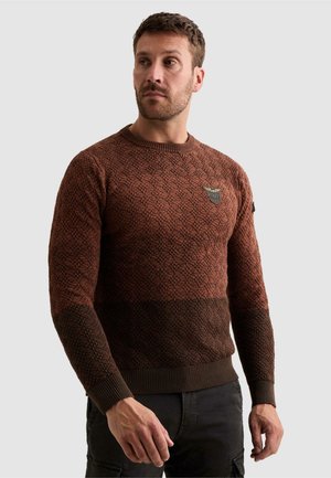 Homme portant un pull en maille marron à motif avec un patch PME sur la poitrine, regardant sur le côté sur un fond clair.