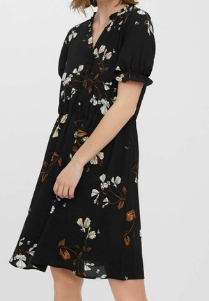 Frau trägt ein schwarzes knielanges Kleid mit weißem und braunem Blumenmuster, kurzen Puffärmeln und einem gerafften Taillenbund.