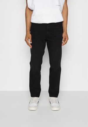 Mann trägt schwarze Jeans mit geradem Bein, weiße Sneaker und ein weißes Sweatshirt mit Fronttasche, steht vor einem einfachen Hintergrund.