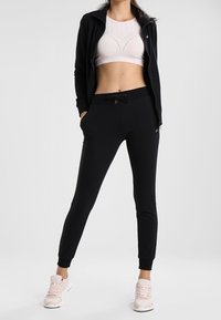 Zwarte jas met rits over een lichtroze cropped top, gecombineerd met zwarte joggerbroeken en lichtroze sportschoenen. Gladde stof, nauwsluitend ontwerp.