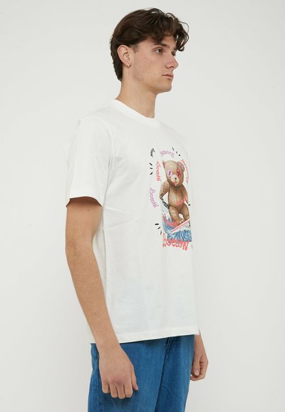 Giovane uomo con capelli castani corti che indossa una t-shirt bianca con un orsetto grafico che surfa sulle onde, abbinata a jeans blu.