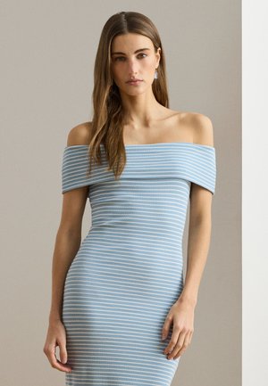 Žena s dlhými hnedými vlasmi, oblečená v svetlomodrých prispôsobených šatách s bielymi vodorovnými pruhmi, stojaca pred jednoduchým pozadím.