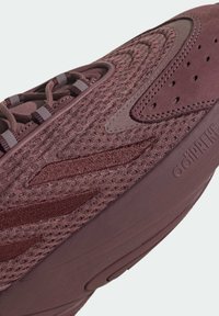 Bordeauxfärgad träningssko med en strukturerad stickad ovandel, släta läderdetaljer och en vadderad sula med en distinkt logotyp på sidan.