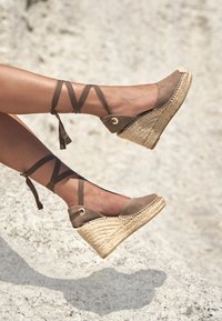 Sandales compensées en daim marron avec une semelle en jute, présentant des brides croisées à la cheville et un design à bout arrondi. Tige texturée et détails tissés.