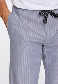 Man in een wit shirt en blauwgestreepte broek met zwarte koord, met zijn linkerhand in de broekzak tegen een witte achtergrond.