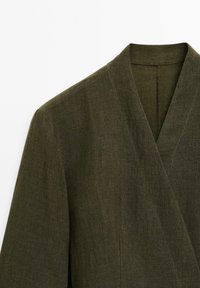 Veste en lin vert olive avec un décolleté en V et un design enveloppant. Présente une texture subtile et des coutures nettes le long des bords.