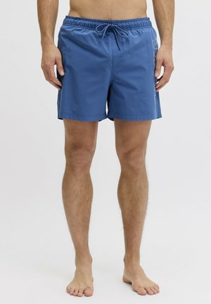 Homme portant un short de bain bleu avec un logo imprimé sur le côté, debout pieds nus sur un fond blanc uni.
