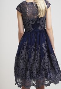 Laivastonsininen mekko hihattomilla hihoilla, jossa on läpikuultava pitsitoppi ja hienostuneet hopeanväriset brodyyrit. Täysihelmainen hame, jossa on aaltoileva pitsireuna.