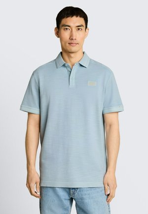 Mann trägt ein hellblaues kurzärmeliges Poloshirt und hellblaue Jeans, steht vor einem schlichten hellgrauen Hintergrund.