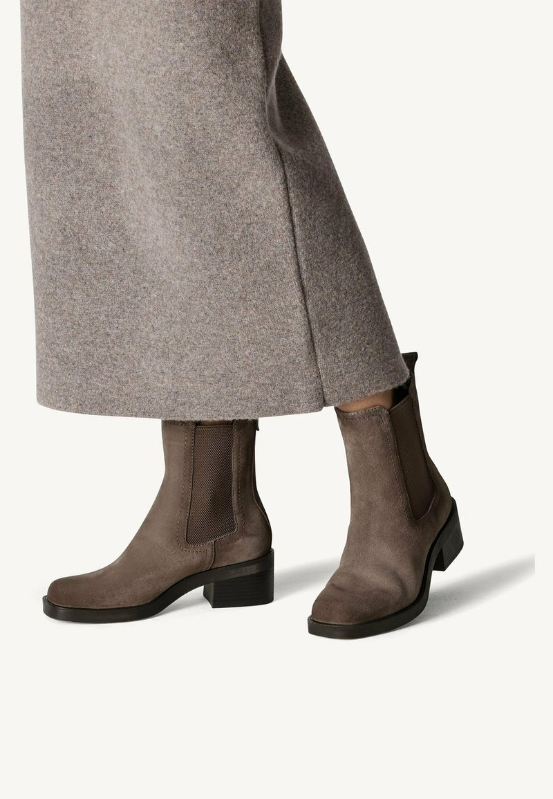 Bottines en daim marron avec des empiècements latéraux élastiques, un bout arrondi et un petit talon bloc, portées avec une longue jupe texturée grise.