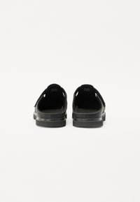 Zapatos negros tipo slip-on con parte superior texturizada, correa de velcro ajustable y suela de goma resistente. Diseño minimalista y funcional.
