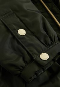 Tela de nylon negra con puños fruncidos, que presenta dos botones a presión plateados en la manga. Textura suave con detalles de costura visibles.