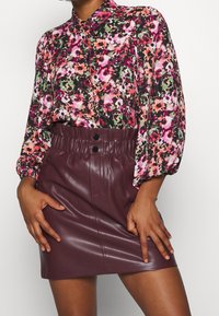 Blouse fleurie avec des motifs roses, verts et noirs ; manches longues. Jupe en simili cuir bordeaux avec ceinture élastique et deux boutons noirs.