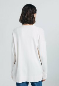 Bílý pletený svetr s volným střihem, dlouhými rukávy a žebrovaným zakončením. Má klasický kulatý výstřih a jednoduchou, hladkou texturu.