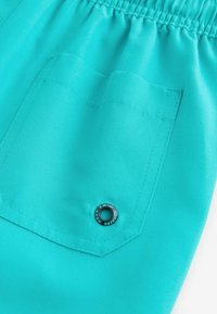 Pantaloni da bagno turchesi con cintura testurizzata, una tasca posteriore con occhiello metallico decorativo e finitura in tessuto liscia.