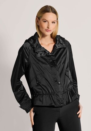 Frau trägt eine schwarze glänzende Kapuzenjacke mit Fronttaschen und elastischem Taillenbund, posiert mit einer Hand auf der Hüfte vor hellem Hintergrund.