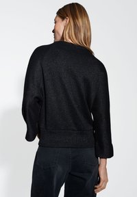 Pull noir avec une finition métallique texturée. Comprend des manches trois-quarts et une coupe décontractée avec un ourlet en côtes. Porté avec un pantalon sombre.