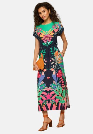 Vrouw draagt een kleurrijke, bloemige maxijurk met toekan- en sterrenpatronen, houdt een oranje geweven clutch vast en draagt oranje sandalen met bandjes.