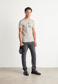 Pelēks kokvilnas t-krekls ar melno "Calvin Klein Jeans" logotipu, apvienots ar pelēkām džinsiem un melnām kurpēm, tur mazus melnus maisiņu.