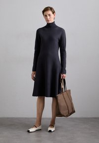 Donkergrijze gebreide jurk met een hoge halslijn, lange mouwen en een uitlopende zoom; gecombineerd met een beige draagtas en lichte schoenen met zwarte puntjes.