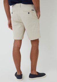 Pantalones cortos de algodón beige con corte recto, que cuentan con bolsillos traseros con botones y bolsillos laterales, combinados con zapatos oscuros tipo slip-on.