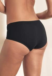 Juodos vientisos hipster stiliaus apatinės kelnaitės su lygiu paviršiumi, žemu liemeniu ir subtiliais siūlėmis palei kraštus.