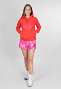 Roter Kapuzenpullover mit Logo, kombiniert mit einem plissierten rosa Rock mit Camouflage-Muster. Weiße Sneakers runden das Outfit ab.