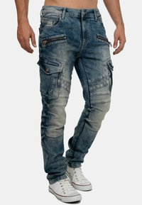 Pantaloni cargo in denim azzurro chiaro con numerose tasche con cerniera, zone sbiadite e un design slim fit, abbinati a sneakers bianche.