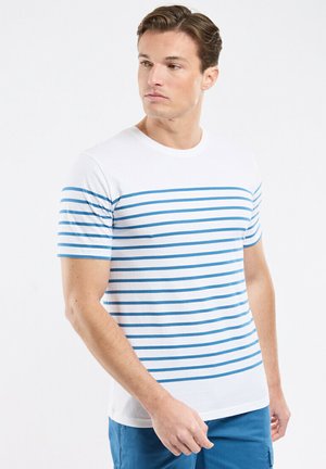 Homme portant une chemise blanche à manches courtes avec des rayures horizontales bleues, regardant vers sa gauche, associé à un pantalon bleu sur un fond blanc uni.