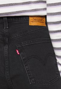 Svarta denimjeans med en orange läderlapp, röd logotagg och kurvad söm vid bakfickan. Randig topp delvis synlig.