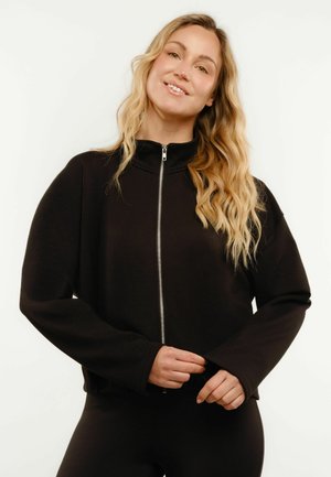 Femme souriante aux longs cheveux blonds ondulés portant une veste noire zippée à manches longues, debout devant un fond clair uni.