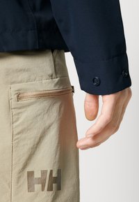 Chaqueta de marina con puño abotonado, combinada con pantalones caqui que tienen un bolsillo con cremallera y un logo "HH" marrón en la pierna.