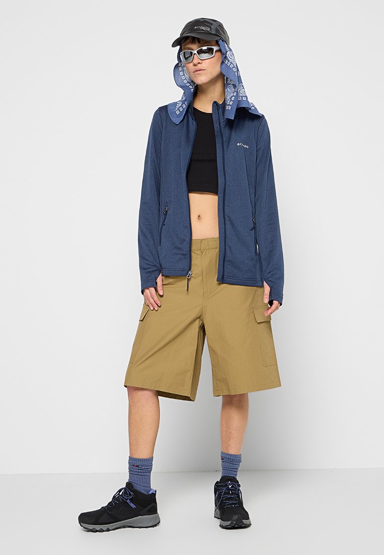 Blauwe ritsjack over een zwarte crop top, tan cargo shorts, zwarte schoenen, blauwe sokken, een zonnebril en een zwarte pet met een bandana.