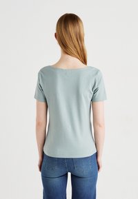 Tricou scurt, de culoare albastru deschis, dintr-un material moale, cu mâneci scurte și un decolteu larg, având o formă ajustată și o textura netedă. Vedere din spate care arată designul umerilor.