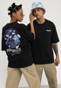 Zwei Models tragen schwarze "MEMORIZE" T-Shirts, eines zeigt ein Koi-Fisch-Design auf dem Rücken, kombiniert mit beigen Cargohosen und Accessoires.