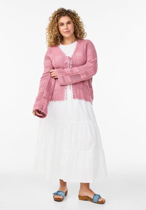 Kobieta z kręconymi włosami, ubrana w różowy dziany sweter na guziki, nałożony na białą, warstwową sukienkę oraz niebieskie drewniane sandały na platformie z odkrytymi palcami.