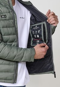 Izolēta olīvu zaļas krāsas jaka ar rāvējslēdzēju priekšpusē, aprīkota ar melnu iekšējo odere un kabatiņu, kurā atrodas kopšanas instrukcijas. Zem jakas redzama balta T-kreklu.
