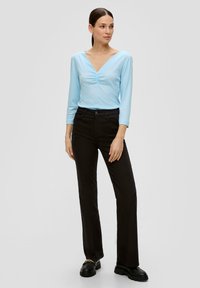 Helles blaues Langarmoberteil mit V-Ausschnitt und gerafftem Detail, kombiniert mit hochgeschnittenen schwarzen Hosen und klobigen schwarzen Schuhen.