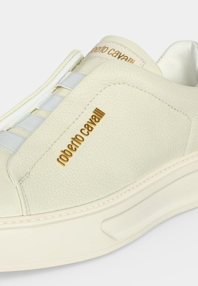 Gros plan d'une sneaker en cuir couleur crème avec des lacets élastiques blancs et le logo doré "roberto cavalli" sur le côté.