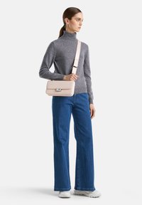 Donna che indossa un dolcevita grigio, jeans a gamba larga blu, sneaker bianche e una borsa a tracolla rosa chiaro con una larga tracolla sulla spalla.