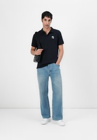 Mornarsko modra polo majica s kratkimi rokavi in kontrastnim logotipom, v kombinaciji s svetlo modrimi širokimi hlačami in belimi supergami. Vključena je torbica preko ramena.