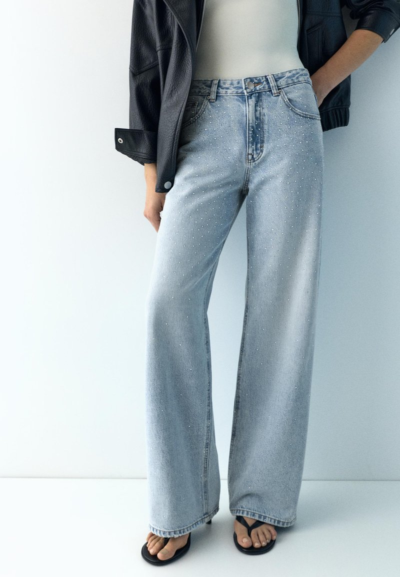 Kvinde iført lyseblå jeans med vide ben og små rhinsten, sort læderjakke, hvid top og sorte flip-flop sandaler.