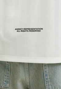 Camisa blanca con rayas verticales, que presenta una impresión negra que dice "REPRESENTACI�ÓN DE AGENCIA TODOS LOS DERECHOS RESERVADOS" cerca del dobladillo. Llevada con jeans de color azul claro.