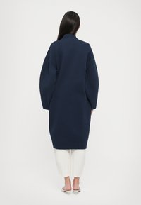 Manteau oversize bleu marine avec un col haut, des épaules tombantes et des manches longues. Tissu doux et silhouette mi-longue, associé à un pantalon blanc.