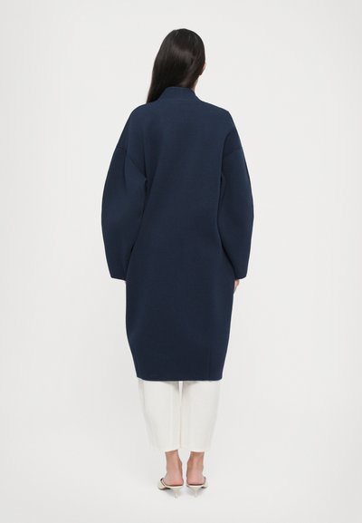 Tumši zilas krāsas lielais mētelis ar augstu apkakli, kritušām plecu līnijām un garām piedurknēm. Mīksts audums un ceļgala garuma siluets, apvienots ar baltām biksēm.