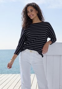 Frau in einem schwarz-weiß gestreiften Pullover und weißen Hosen lächelt, steht auf einer Holzpier am ruhigen Ozean unter klarem Himmel.