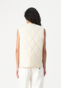 Quiltet vest i kremfarge, med en bølgete mønster, ermeløs design og subtile sømdetaljer. Sett fra baksiden.
