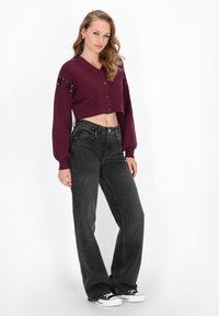 Cárdigan burdeos recortado con botones y cordones en las mangas, combinado con jeans de mezclilla negros de cintura alta y sneakers negros.