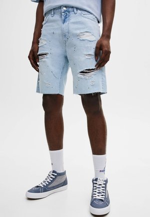 Homme portant un short en jean déchiré bleu clair, des chaussettes blanches et des baskets montantes bleues, debout devant un fond uni.